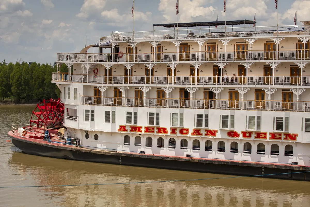 The American Queen moored on the river, red stern paddle wheel beside tiered white balconies and her name in red