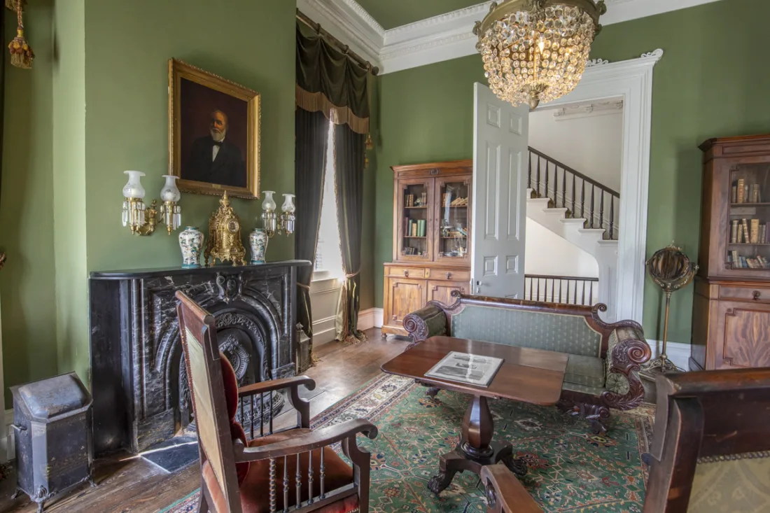 A green walled parlour with marble fireplace, gilt clock, chandelier and a portrait above the mantel