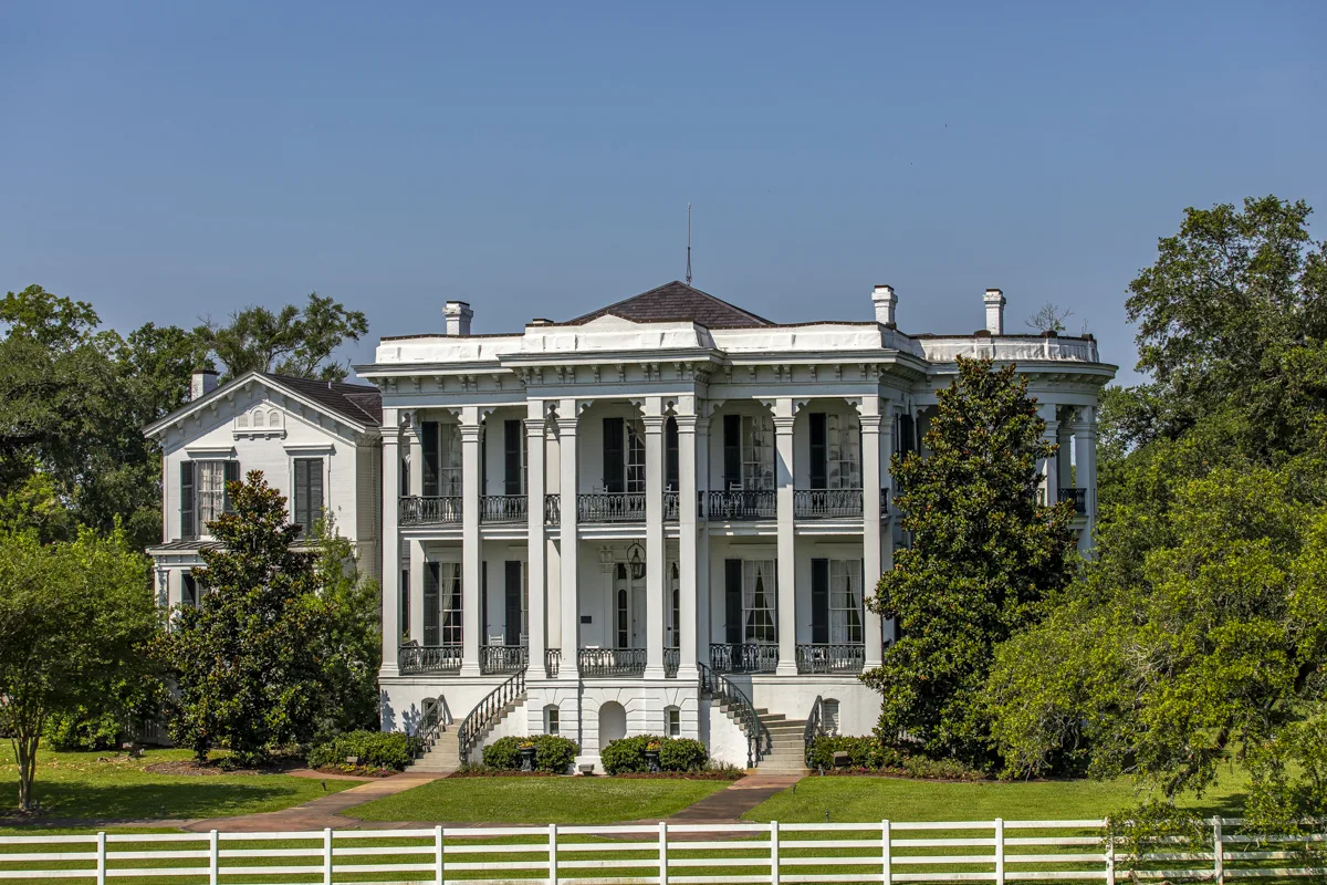 A white columned antebellum mansion behind a low white fence and magnolias on the Louisiana riverbank
